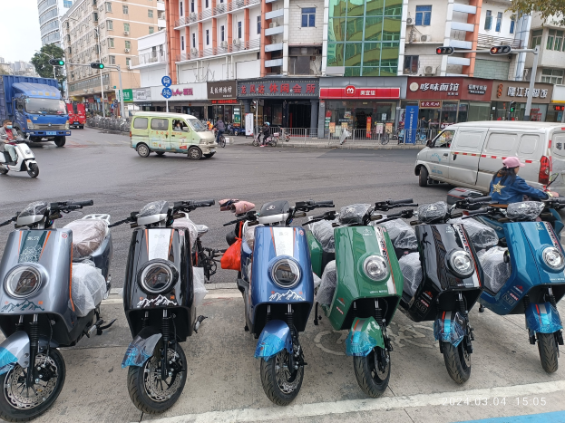 Eine Reihe von Elektro-Scootern, die entlang einer Straße geparkt sind, mit einer Person in einer Mütze davor, Gebäuden, Schildern, Verkehrszeichen, Bäumen und einem klaren blauen Himmel im Hintergrund.