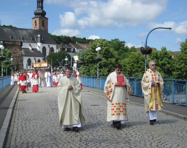 Eine Gruppe von Priestern, die eine Straße mit Laternen, Geländern und Bäumen entlanggehen; im Hintergrund sind Gebäude und ein bewölkter Himmel zu sehen; einer der Priester hält ein Buch in der Hand.