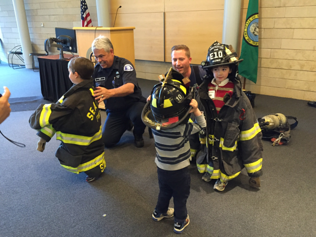 Eine Gruppe von Kindern in Feuerwehruniform mit einem jungen Jungen in Uniform, der einen Helm hält, einer Person mit einer Kamera links daneben und Hintergrundelementen wie Stühlen, einem Tisch, einem Podium, einem Mikrofon, einer Fahne und einer Wand.