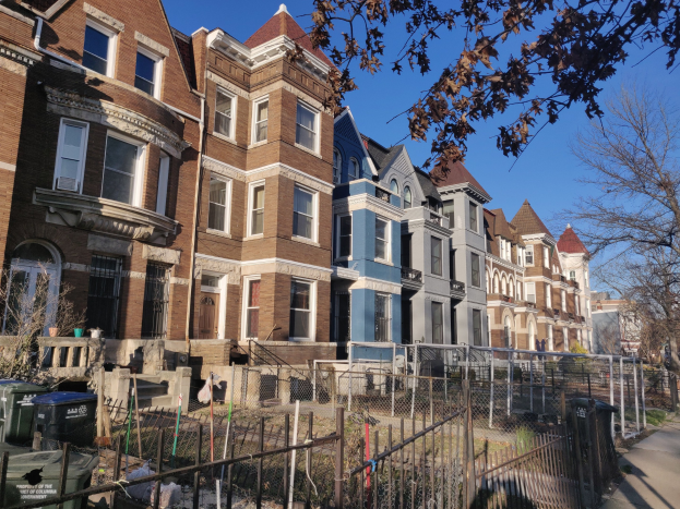 Reihe von H├Ąusern in einer Wohngegend mit einem Zaun, Fenstern, B├Ąumen, Mülltonnen und sichtbarem Himmel im Hintergrund.