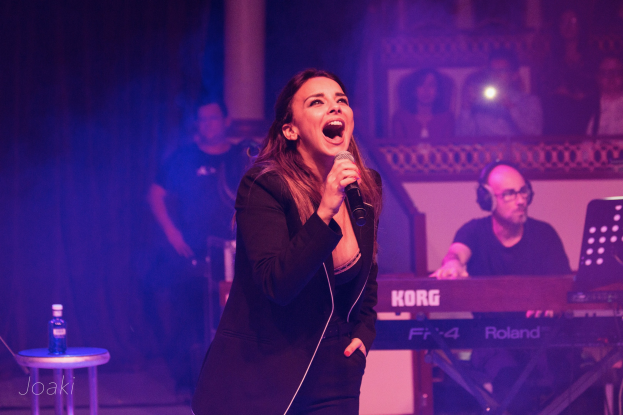 Eine Frau singt in ein Mikrofon auf einer Bühne, rechts neben ihr ein Pianist und einige Menschen im Hintergrund, links auf einem Hocker eine Flasche.