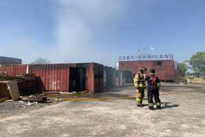 Zwei Feuerwehrleute in Helmen vor einem qualmenden Gebäude mit einem Namensschild, umgeben von verstreuten Containern und Holzgegenständen, mit Bäumen und einem klaren blauen Himmel im Hintergrund.