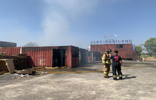 Zwei Feuerwehrleute in Helmen vor einem qualmenden Gebäude mit einem Namensschild, umgeben von verstreuten Containern und Holzgegenständen, mit Bäumen und einem klaren blauen Himmel im Hintergrund.