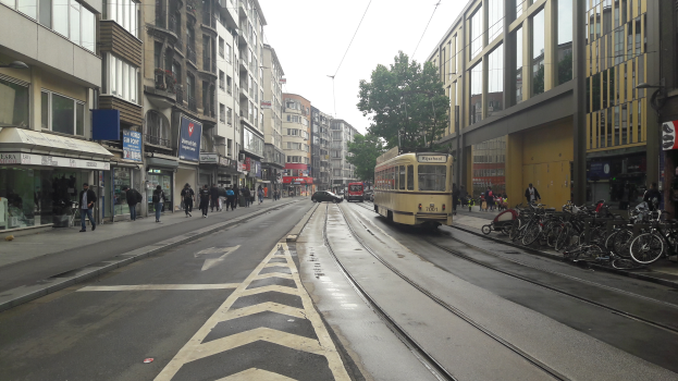 Eine Stadtstraße mit einer gelben Tram, Gebäuden mit Fenstern, Schildern, Fußgängern, parkenden Fahrrädern, einer Straßenlaterne, Drähten, einem Baum und einem bewölkten Himmel.
