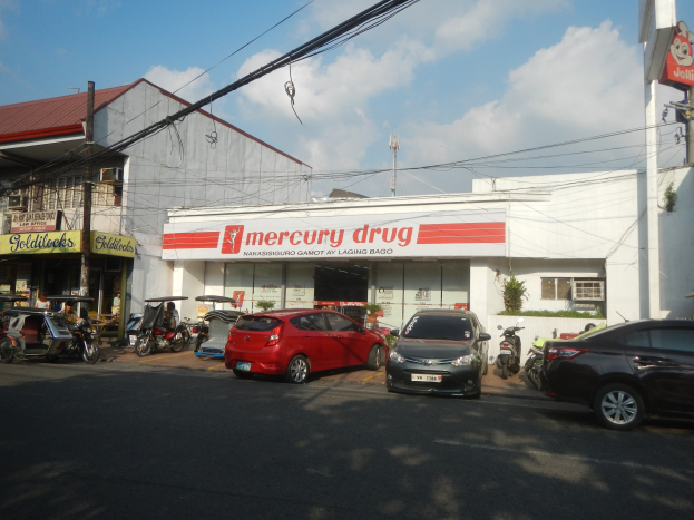 Eine belebte Straßenszene mit Fahrzeugen, Gebäuden mit Schildern, Strommasten mit Drähten, einem bewölkten Himmel und einer Mercury Drug Apotheke mit parkenden Autos davor.