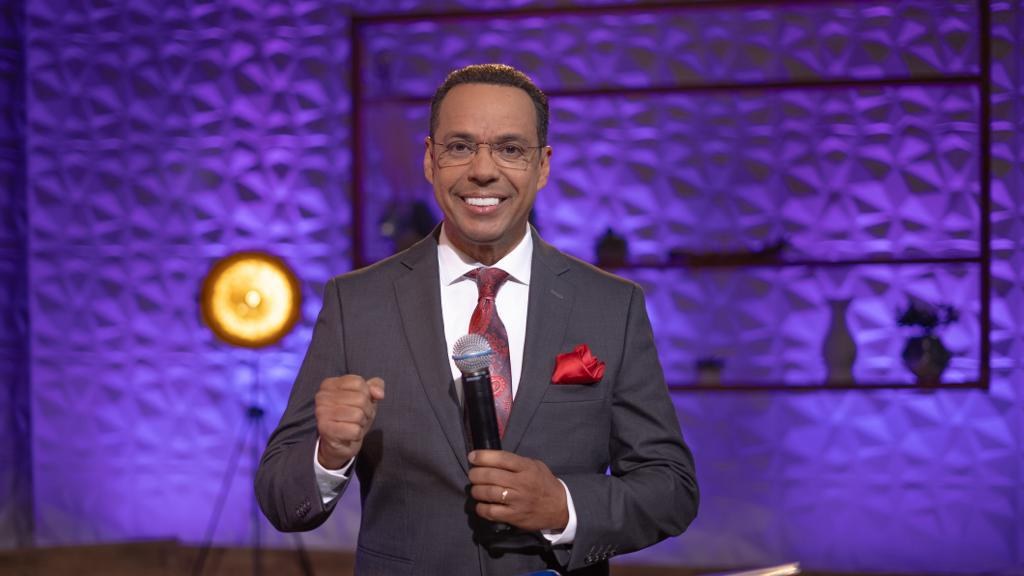 Stephen Colbert, Moderator von *The Daily Show with Trevor Noah*, steht auf einer Bühne und hält ein Mikrofon in der Hand, trägt einen Anzug, Krawatte und Brille, mit einer Lampe und Ständer im Hintergrund.