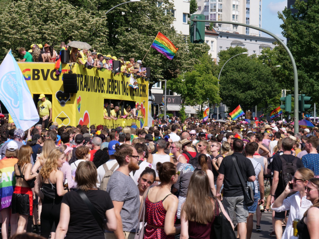 Eine große Menge von Menschen mit Mützen und Fahnen geht eine Straße in Berlin, Deutschland, entlang, mit einem gelben Lastwagen, Laternenmasten und Verkehrszeichen im Hintergrund, flankiert von Bäumen und Gebäuden unter einem bewölktem Himmel.