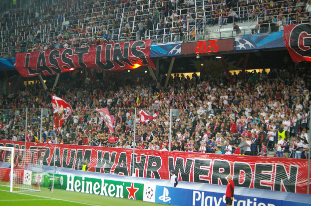 Ein Fußballspiel in einem Stadion mit einer großen Menge, saftig grünem Rasen, einem Torpfosten auf der linken Seite, Bodenbanner, Menschen mit Fahnen in der Hand und Hintergrundgeländer, Tafeln, Lampen und andere Gegenstände.