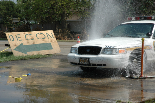 Ein Streifenwagen fährt durch eine Wasserlache, mit einem Schild, einer Verkehrskegel und anderen Gegenständen auf dem Boden, mit Bäumen, Gebäuden und einem klaren blauen Himmel im Hintergrund.