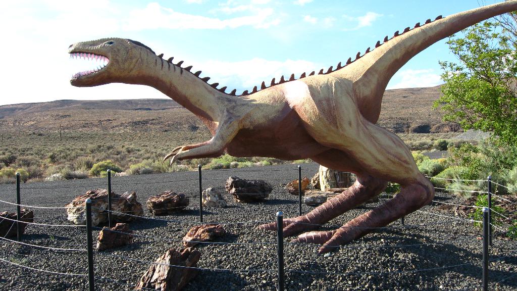 Eine Dinosaurierstatue steht mit einigen Gegenständen auf dem Boden, mit Bäumen und Himmel im Hintergrund.