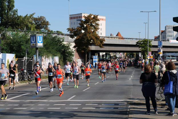 Gruppe von Läufern bei einem Marathon auf einer Straße mit Bäumen, Laternen, Schildern, Fahrrädern, einem Zaun, Gras, einer Brücke, Gebäuden und einem klaren blauen Himmel.