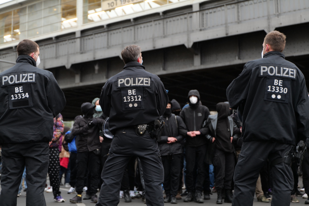 Polizisten in schwarzer Uniform und Masken stehen vor einer Menge während einer Demonstration, mit einer Brücke und einem Gebäude im Hintergrund.