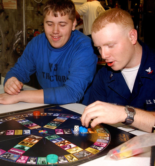 Zwei Männer sitzen an einem Tisch und spielen ein Brettspiel mit Würfeln und anderen Gegenständen auf dem Tisch, und ein paar Menschen stehen im Hintergrund.