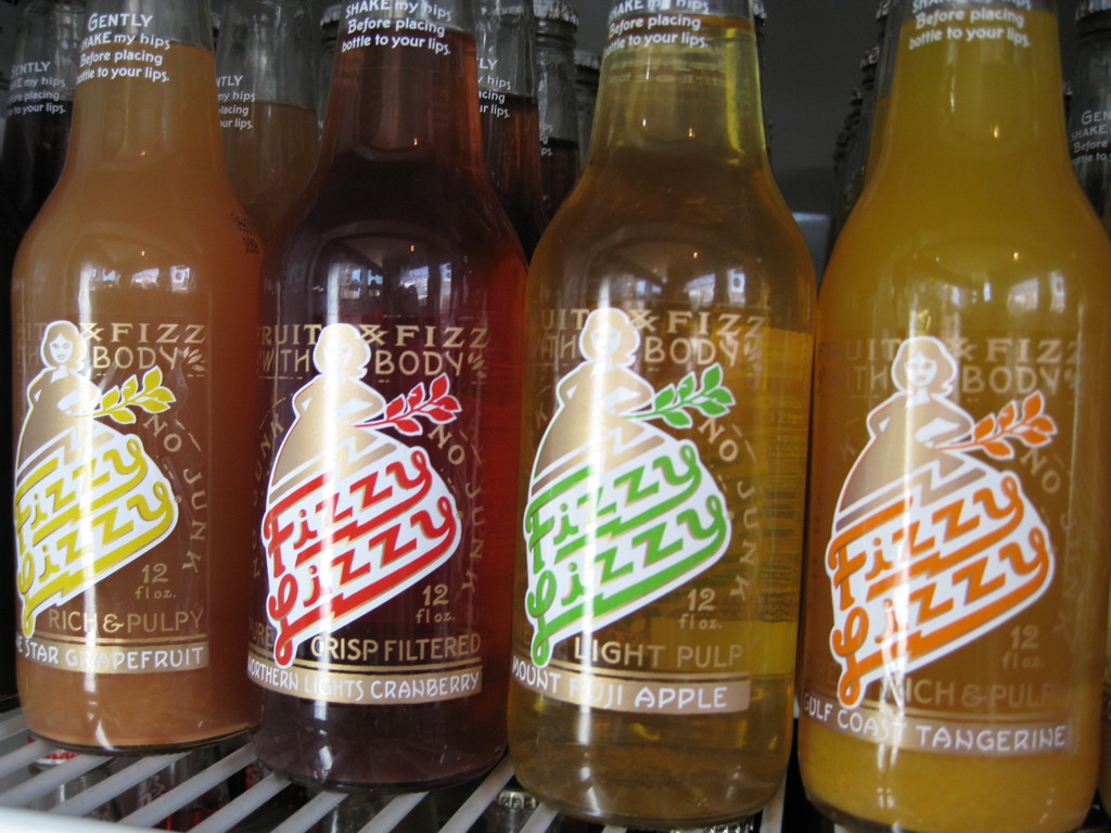 Drei farbenfrohe Getänkeflaschen mit der Aufschrift 'Fizzy Lizzy' in Gelb, Rot und Orange auf einem Tisch