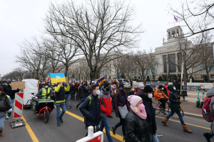 Ein großer Protestmarsch mit Menschen, die eine Straße entlang gehen, einige halten Schilder und andere fahren Fahrräder, mit Bäumen und einem klaren blauen Himmel im Hintergrund.