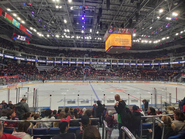 Eine große Menschenmenge in einer Hockeyarena, einige sitzen und einige stehen, schauen ein Spiel mit hellen Lichtern und Bannern an der Decke.