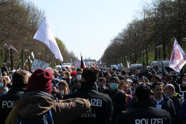 Eine große Gruppe von Menschen steht vor einer Menge von Polizeibeamten, einige tragen Mützen und Masken, während sie auf einer Demonstration in Berlin, Deutschland, mit Schildern, Fahnen, Laternenmasten, Bäumen, Fahrzeugen, einem Gebäude und dem Himmel im Hintergrund demonstrieren.