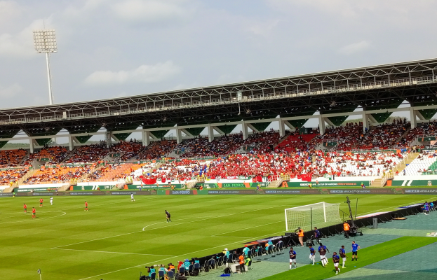 Ein Fußballspiel in einem großen Stadion mit Spielern auf dem Boden, einem Tor, einem Zaun, einer Fahne mit Text, Metallrahmen, einem Dach mit Deckenleuchten und einem bewölkten Himmel.