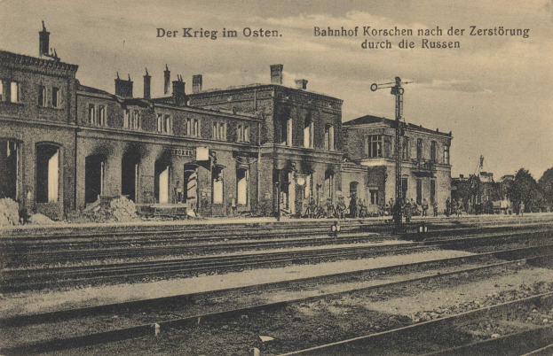 Schwarz-weiß-Fotografie eines Bahnhofs in Korschen Ost, Deutschland, mit Bahnschienen, Gebäuden, Strommasten, Menschen, Bäumen und Himmel.