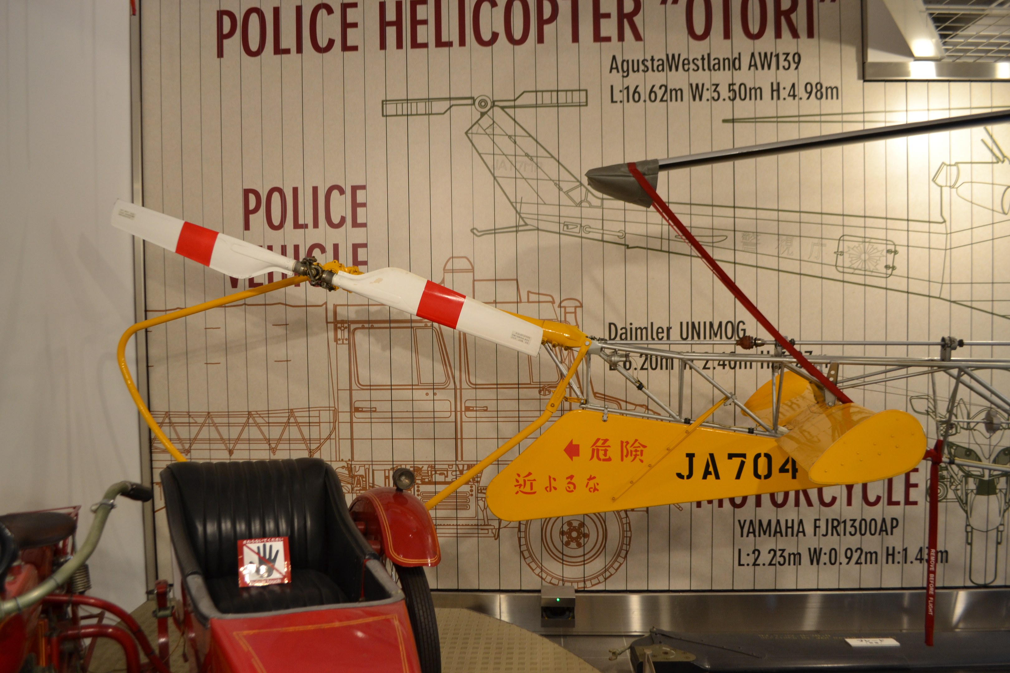 Polizeihubschrauber in einem Museum ausgestellt, mit einem Fahrzeug links und einer Informationswand im Hintergrund, beleuchtet von Deckenlampen.