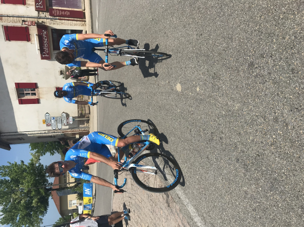 Eine Gruppe von Radfahrern fährt eine Straße entlang an einem Gebäude mit vernagelten Fenstern und Text vorbei, Bäumen, einem blauen Himmel und Zuschauern auf der Straße darunter.
