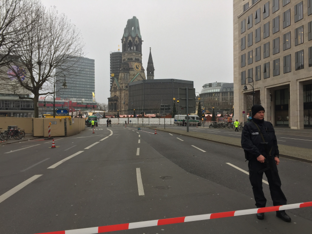 Ein Polizist in schwarzer Uniform und Mütze steht auf einer Straße in Berlin umgeben von Absperrbaken, Fahrrädern, Laternenmasten, Bäumen und Gebäuden, im Hintergrund ist ein Kirchturm und ein klarer blauer Himmel zu sehen.