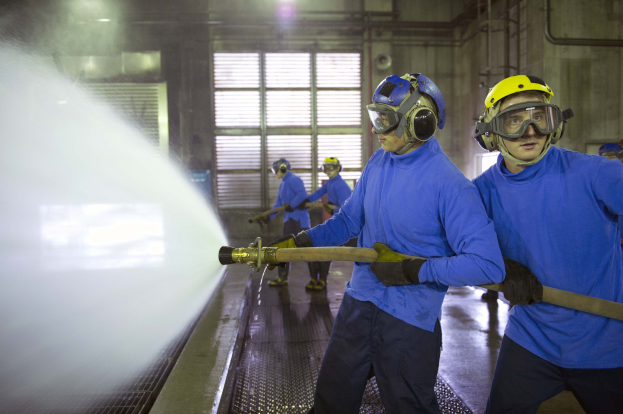 Gruppe von Männern in blauen Hemden und gelben Helmen, die an einer Maschine arbeiten, wobei einer einen Schlauch hält und Wasser auf den Boden sprüht, in einer Fabrikumgebung.
