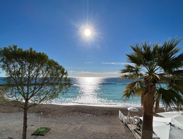 Ein sonniger Strand mit Palmen, Schirmen und üppiger Vegetation unter einem strahlend blauen Himmel an der französischen Riviera.