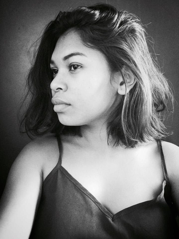 Eine selbstbewusste Frau in einem schwarzen Kleid posiert in einem Schwarz-Weiß-Foto, ihr glatter Bob und natürliches Make-up unterstreichen ihre selbstbewusste Haltung an einer Wand.