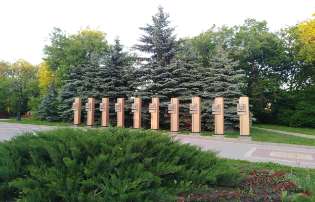 Ein Park mit einer Reihe von Bäumen in der Mitte, umgeben von blühenden Pflanzen, Gras, einem Weg, einem Straßenpfahl, einer Gedenktafel für Opfer des Tschernobyl-Unfalls, einer Gruppe von Bäumen und einem bewölkten Himmel.