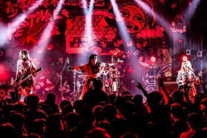 Ein Live-Auftritt einer japanischen Rockband mit Gitarristen und Sängern auf der Bühne, Mikrofone und Ständer, einer beleuchteten Wandmalerei im Hintergrund und einem Publikum darunter.