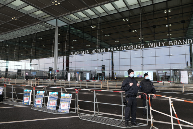 Zwei Polizisten in schwarzen Uniformen und Mützen stehen vor einem großen Glasgebäude mit Text darauf, mit Absperrungen und Texttafeln im Vordergrund.