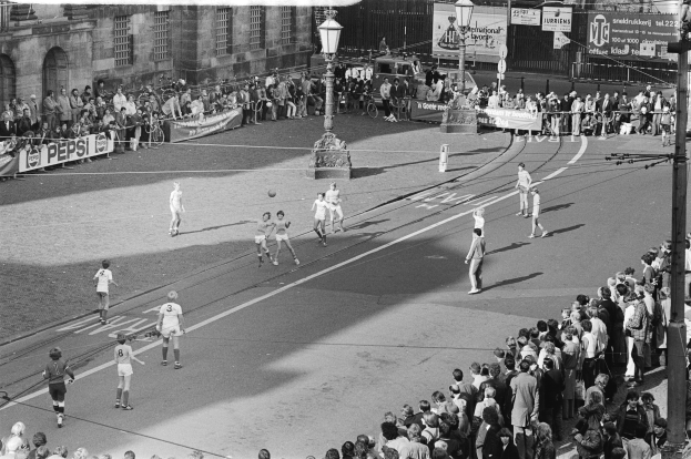 Schwarzes Foto eines auf der Straße ausgetragenen Fußballspiels mit Zuschauern am Straßenrand, Laternenpfählen und Gebäuden mit Fenstern.