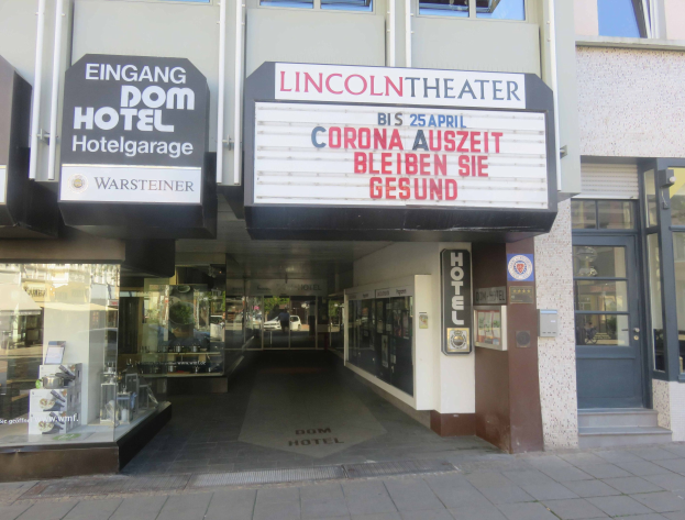 Außenansicht des Lincoln Theaters in Berlin, Deutschland, mit Glasfenstern und -türen sowie einer Schautafel und einem belebten Innenraum, der durch den Eingang sichtbar ist.