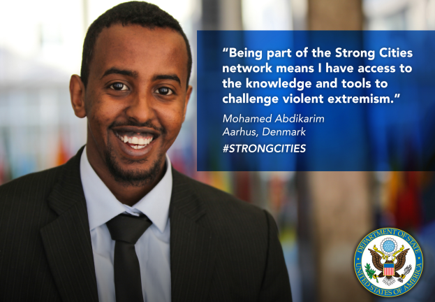 Ein Mann im Anzug und Krawatte, der strahlend in die Kamera lächelt, mit einem Logo und Text auf der rechten Seite, der besagt "Teil des starken Städtenetzwerks zu sein bedeutet, Zugang zu Wissen und Werkzeugen zu haben, um gewalttätigen Extremismus in Frage zu stellen", und einem unscharfen Hintergrund.