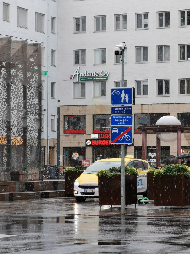 Gelbes Taxi fährt auf regennasser Stadtstraße mit einer Person mit Schirm, einem Schild, blühenden Pflanzen und einem Gebäude mit Fenstern im Hintergrund.