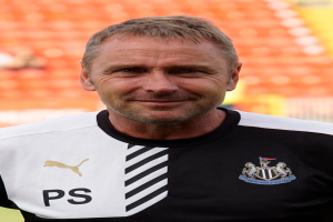 Ein Mann in einem schwarzen und weißen T-Shirt steht selbstbewusst auf einem Fußballfeld und lächelt entschlossen, während der Hintergrund unscharf ist.