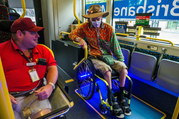 Zwei Männer im Rollstuhl in einem Bus, einer mit rotem T-Shirt und Kappe, der andere mit orangefarbenem Hemd und brauner Mütze, beide mit Masken und Ausweisen, mit gelben Stangen und Glasfenstern, die Bäume, Gebäude und den Himmel zeigen.