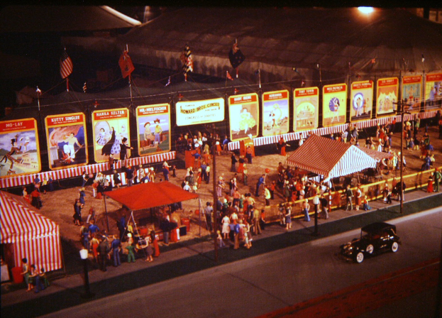 Ein altes nächtliches Karnevalsszenario mit Menschen, einem Auto, Pfählen mit Fahnen, einem Zaun, einem Zelt, Brettern mit Bildern und Text sowie einer Straßenlaterne.
