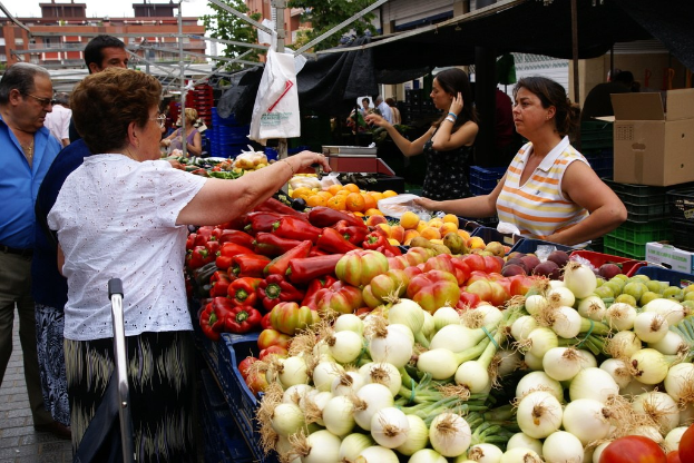 Gemüse in Kunststoffbehältern, eine Gruppe von Menschen, die stehen, Gebäude, Bäume, eine Kartonbox und der Himmel.