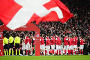 Gruppe von Menschen auf einem Fußballfeld mit einer roten und weißen Flagge im Vordergrund, einem Bogen mit "Bayern Munich vs Bayern Munich Betting Tips & Preview" im Hintergrund und einer großen Menge im Stadion.