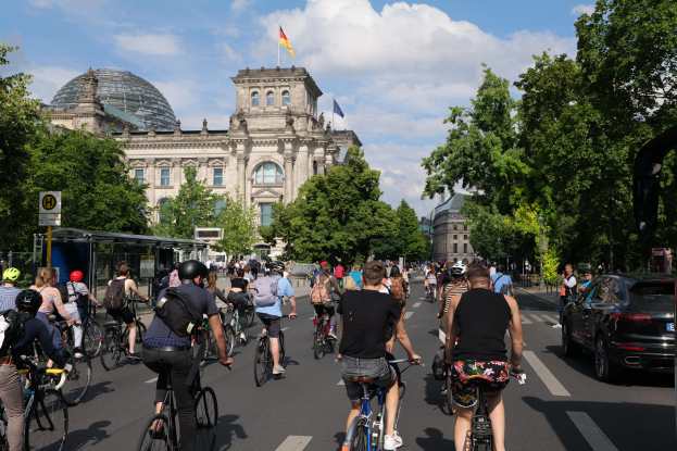 Eine Gruppe von Menschen, die Fahrräder auf einer von Bäumen gesäumten Straße in Berlin, Deutschland, fahren, mit Gebäuden, einer Bushaltestelle auf der rechten Seite und einer Flagge, die auf einem der Gebäude weht, unter einem bewölkten Himmel.