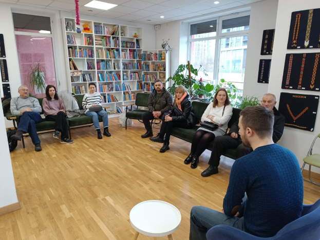 Eine Gruppe von Menschen sitzt um einen Tisch in einem Raum mit Bücherregalen, Pflanzen und einem Fenster, die sich während eines Workshops zur Gesundheit und Wellness in Berlin, Deutschland, unterhalten.