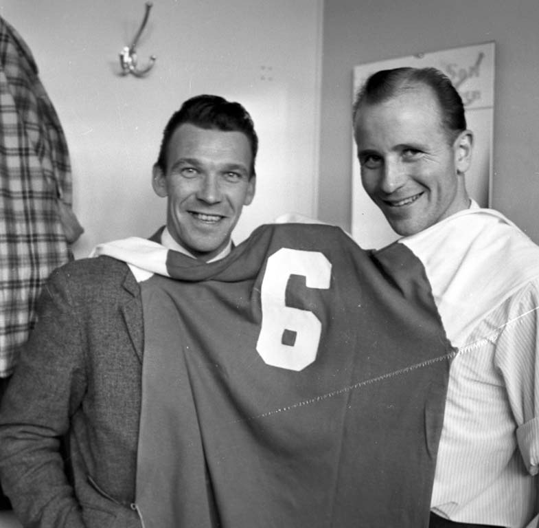 Zwei Männer in Schwarz-Weiß stehen nebeneinander, lächeln und halten ein Fußballtrikot hoch; im Hintergrund ist eine Wand mit einer Tafel und einem Bügel zu sehen.