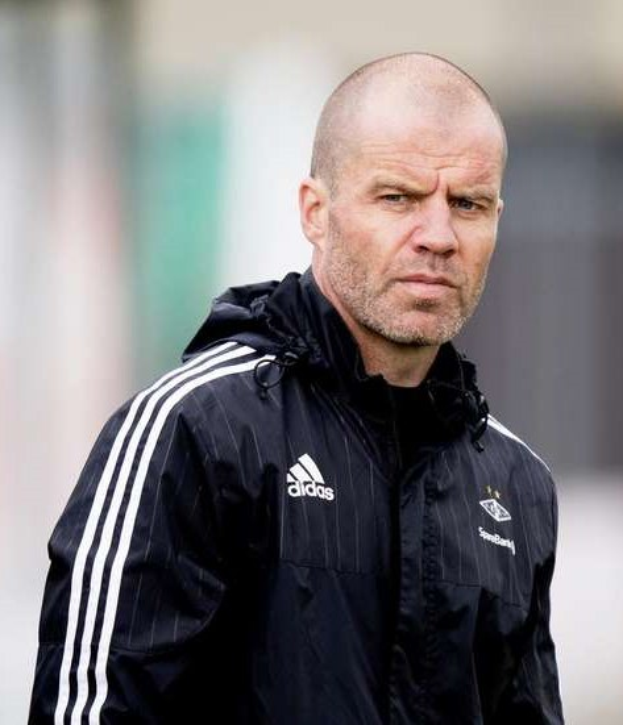 Baldiger Trainer in marineblauer Jacke mit weißen Streifen und weißem Kragen, der auf einem Fußballfeld steht und nachdenklich schaut.