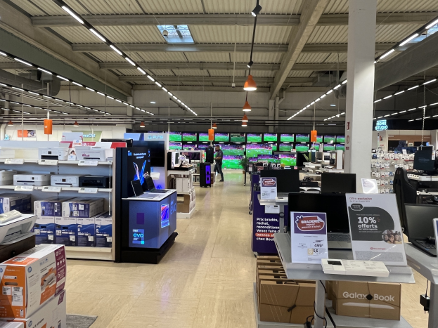 Ein großer Raum in einem Walmart-Laden voller Elektronik wie Computern und Druckern, die in Regalen angeordnet sind, mit Menschen auf dem Boden, einer Säule, Schildern, Deckenlampen und Ventilatoren.