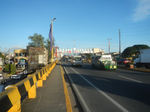 Eine belebte Stadtstraße mit Fahrzeugen, einer Trennung, einem Fahnenmast mit Flagge, Gebäuden, Bäumen, Strommasten mit Drähten und einem bewölkten Himmel im Hintergrund.