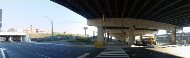 Ein Lastwagen, der unter einer Brücke auf einer Stadtstraße mit Gebäuden, Straßeninfrastruktur und Fahrzeugen unter einem klaren blauen Himmel fährt.