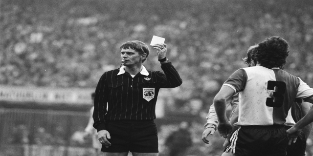 Schwarzes und weißes Bild eines Schiedsrichters, der eine rote Karte vor einem Publikum in Fußballuniformen hält, mit einem Zaun im Hintergrund.