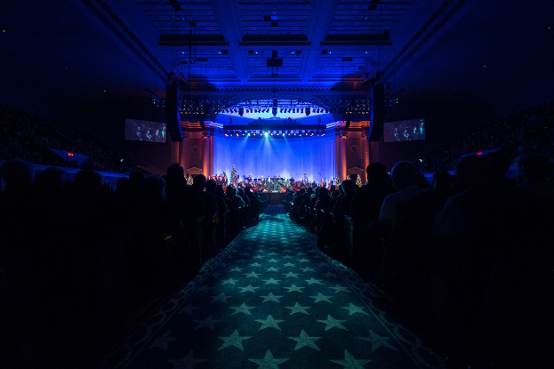 Ein großer Zuschauerraum mit Sitzplätzen, eine Bühne mit Musikern und Deckenbeleuchtung und -fernseher, was auf ein Konzerthaus während der Nationalkonvention 2019 hinweist.